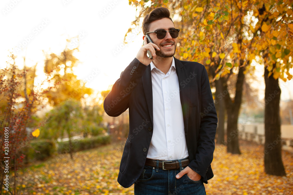 Handsome young stylish male businessman happy and smiling talking on the phone on a walk in the autumn park on the background of yellow leaves