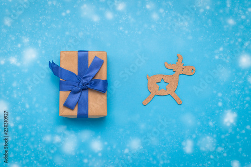 Gift box with a blue ribbon and a Christmas wooden toy Deer on a blue background with the effect of falling snow. The concept of gifts for Christmas and New Year. Flat lay, top view