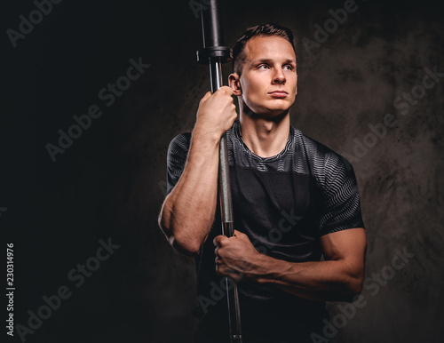 Portrait of a handsome young bodybuilder holding a barbell and looking away on dark background.