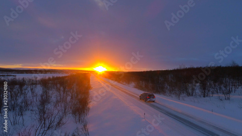 LENS FLARE: Gray tourist car speeds down snowy road at beautiful winter sunset.