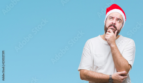 Young caucasian hipster man wearing christmas hat over isolated background with hand on chin thinking about question, pensive expression. Smiling with thoughtful face. Doubt concept.