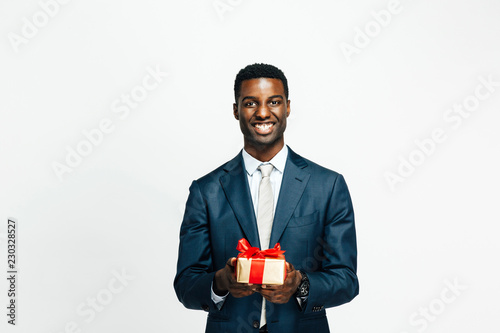 Horizontal portrait of a smiling young man in suit and tie holding a golden gift tied with red ribbon, isolated on white studio background