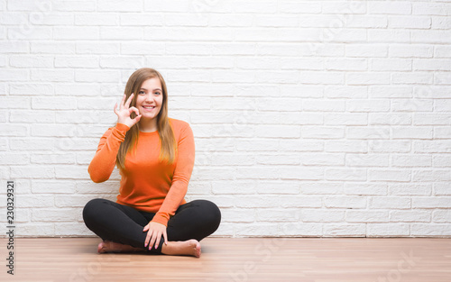 Young adult woman sitting on the floor in autumn over white brick wall doing ok sign with fingers, excellent symbol