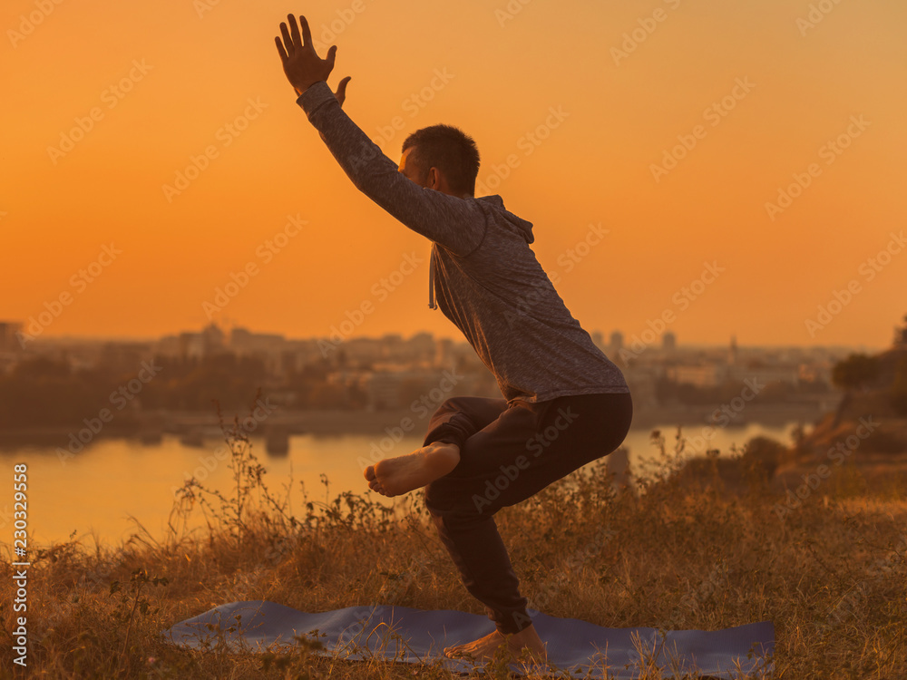 Man doing yoga on sunset with city view, variation of Chair Pose /Utkatasana. 