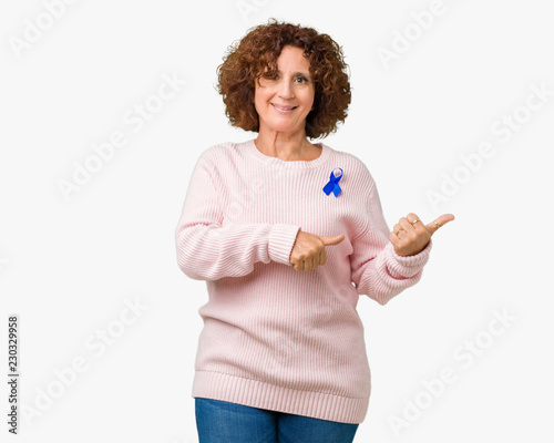 Middle ager senior woman wearing changeable blue color ribbon awareness over isolated background Pointing to the back behind with hand and thumbs up, smiling confident
