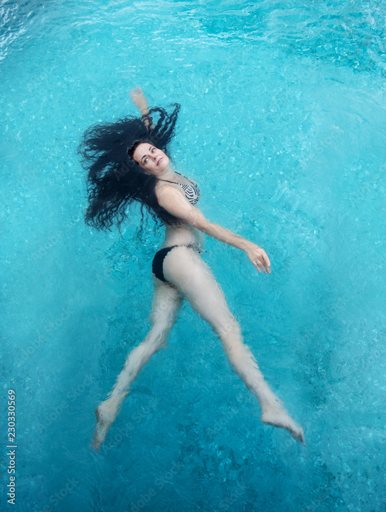 schöne reife Frau im besten Alter mit dunklen Locken im Bikini läuft schwebt laufend schwerelos elegant glücklich schwimmend in türkis blauem Wasser im Pool