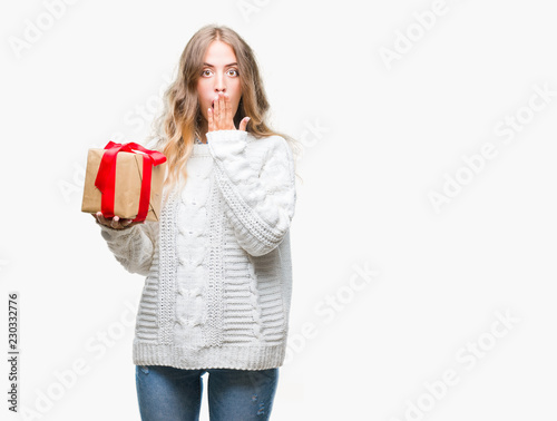 Beautiful young blonde woman holding gift over isolated background cover mouth with hand shocked with shame for mistake, expression of fear, scared in silence, secret concept