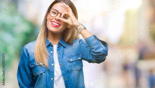 Young beautiful woman over wearing glasses over isolated background doing ok gesture with hand smiling, eye looking through fingers with happy face. © Krakenimages.com
