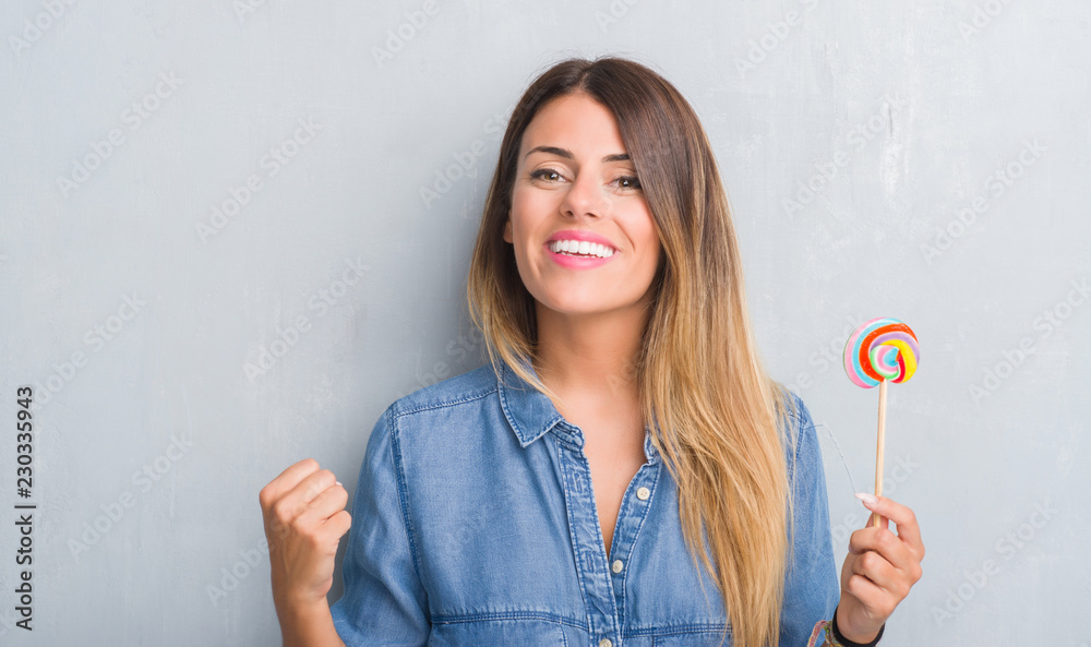 Young adult woman over grey grunge wall eating lollipop candy screaming proud and celebrating victory and success very excited, cheering emotion Young adult woman over grey grunge wall eating lollipop candy screaming proud and celebrating victory and success very excited, cheering emotion