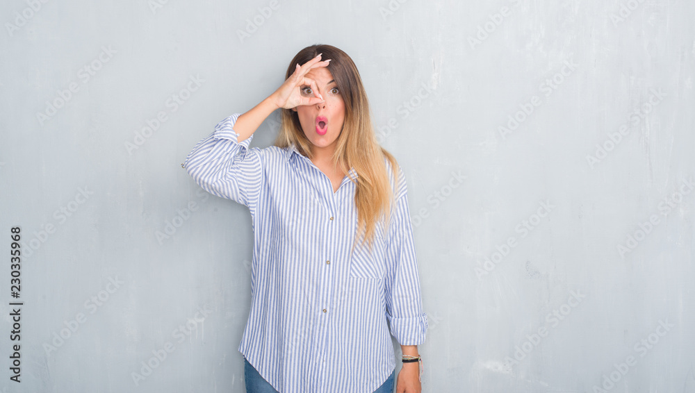 Young adult woman over grey grunge wall wearing fashion business outfit doing ok gesture shocked with surprised face, eye looking through fingers. Unbelieving expression. Young adult woman over grey grunge wall wearing fashion business outfit doing ok gesture shocked with surprised face, eye looking through fingers. Unbelieving expression.