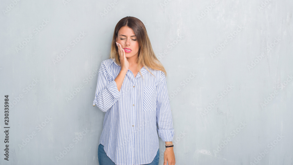 Young adult woman over grey grunge wall wearing fashion business outfit touching mouth with hand with painful expression because of toothache or dental illness on teeth. Dentist concept. Young adult woman over grey grunge wall wearing fashion business outfit touching mouth with hand with painful expression because of toothache or dental illness on teeth. Dentist concept.