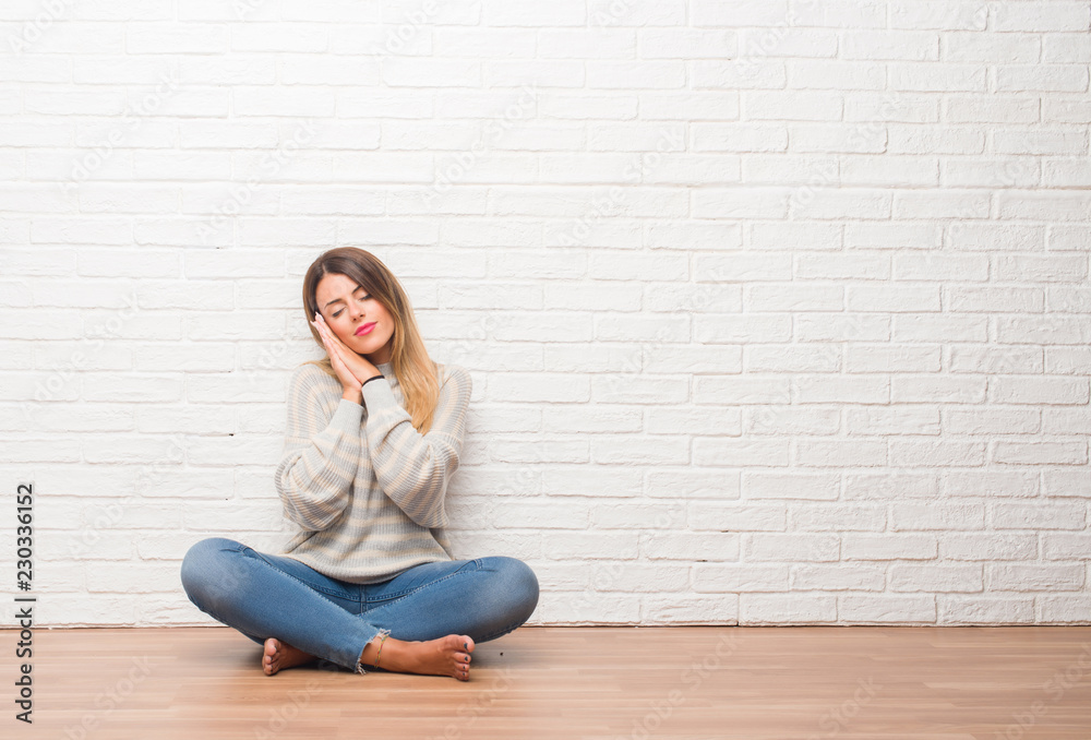 Young adult woman sitting on the floor over white brick wall at home sleeping tired dreaming and posing with hands together while smiling with closed eyes.