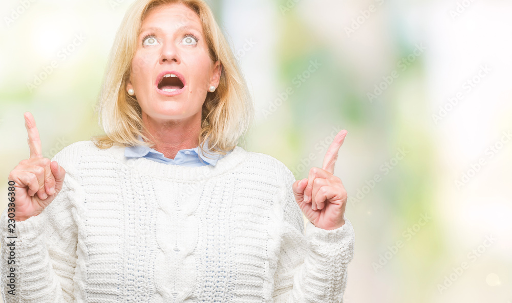 Middle age blonde woman wearing winter sweater over isolated background amazed and surprised looking up and pointing with fingers and raised arms. Middle age blonde woman wearing winter sweater over isolated background amazed and surprised looking up and pointing with fingers and raised arms.