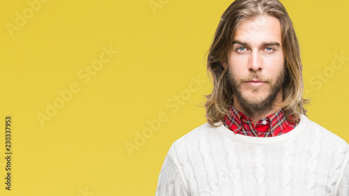 Young handsome man with long hair wearing winter sweater over isolated background with serious expression on face. Simple and natural looking at the camera.