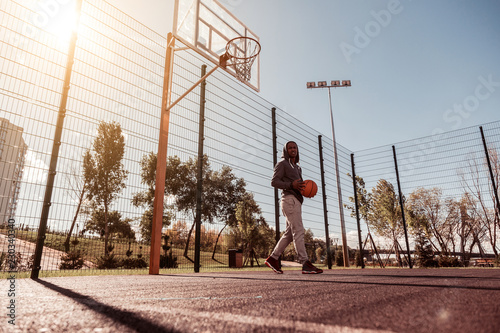 Game territory. Positive joyful man holding a ball while walking in the basketball court