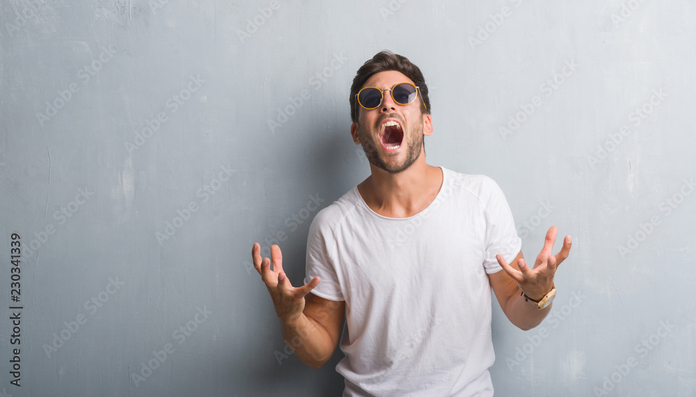 Handsome young man over grey grunge wall wearing sunglasses crazy and mad shouting and yelling with aggressive expression and arms raised. Frustration concept. Handsome young man over grey grunge wall wearing sunglasses crazy and mad shouting and yelling with aggressive expression and arms raised. Frustration concept.