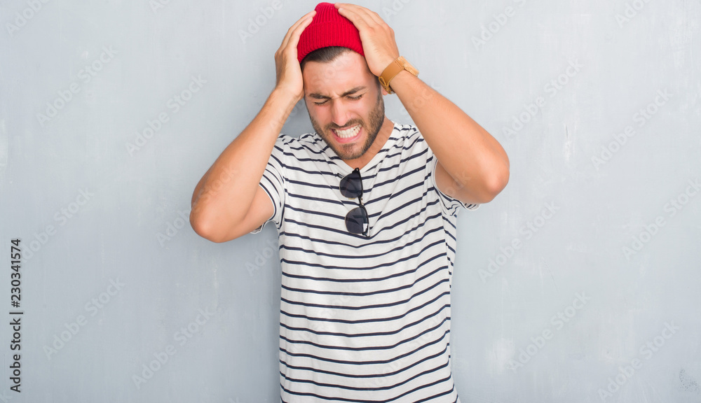 Naklejka premium Handsome young hipster man over grey grunge wall wearing navy t-shirt and wool cap suffering from headache desperate and stressed because pain and migraine. Hands on head.