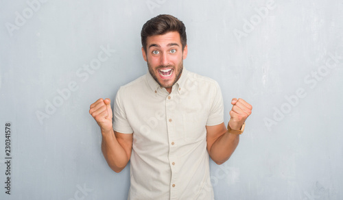 Handsome young man over grey grunge wall wearing summer shirt celebrating surprised and amazed for success with arms raised and open eyes. Winner concept. © Krakenimages.com