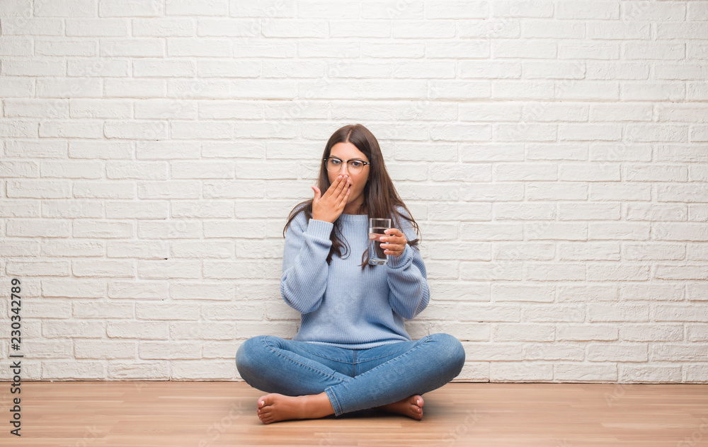 Young brunette woman sitting on the floor drinking glass of water cover mouth with hand shocked with shame for mistake, expression of fear, scared in silence, secret concept