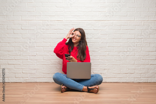 Young brunette woman sitting on the floor using laptop and smartphone with happy face smiling doing ok sign with hand on eye looking through fingers © Krakenimages.com
