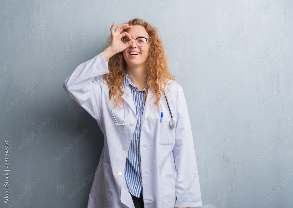 Young redhead doctor woman over grey grunge wall wearing a coat doing ok gesture with hand smiling, eye looking through fingers with happy face. Young redhead doctor woman over grey grunge wall wearing a coat doing ok gesture with hand smiling, eye looking through fingers with happy face.
