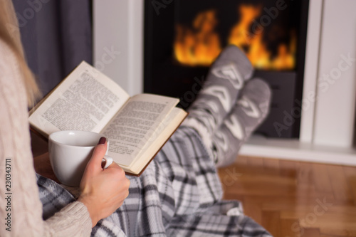 Girl in front of the fireplace reading book and warming feet on fire and legs are covered with blanket, in hand holds cup of hot tea. Winter and cold weather concept at home. Close up, selective focus