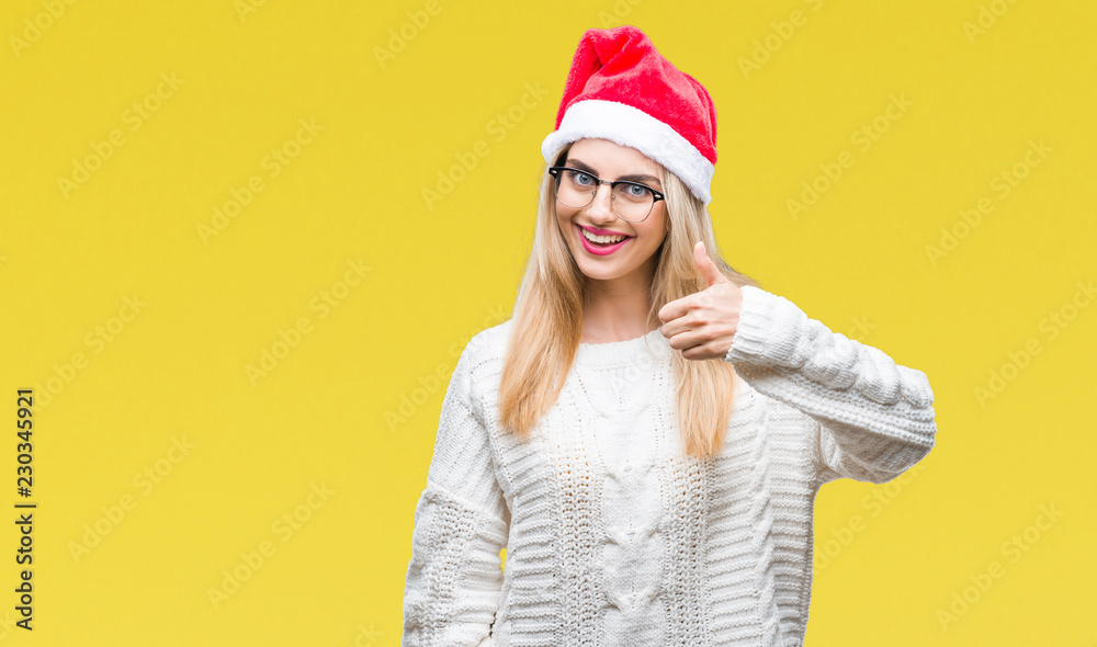 Young beautiful blonde woman wearing christmas hat over isolated background doing happy thumbs up gesture with hand. Approving expression looking at the camera with showing success. Young beautiful blonde woman wearing christmas hat over isolated background doing happy thumbs up gesture with hand. Approving expression looking at the camera with showing success.