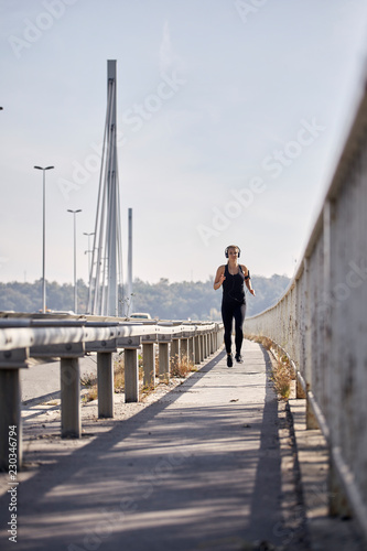 one young woman, running jogging on bridge, cars in background, listening to music on headphones. (smartphone arm holder) sunny Summer day.