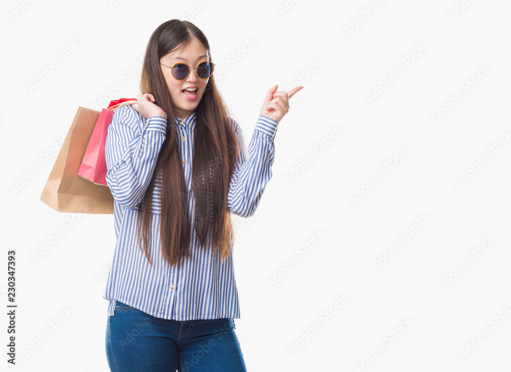 Young Chinese woman over isolated background holding shopping bags on sales very happy pointing with hand and finger to the side Young Chinese woman over isolated background holding shopping bags on sales very happy pointing with hand and finger to the side