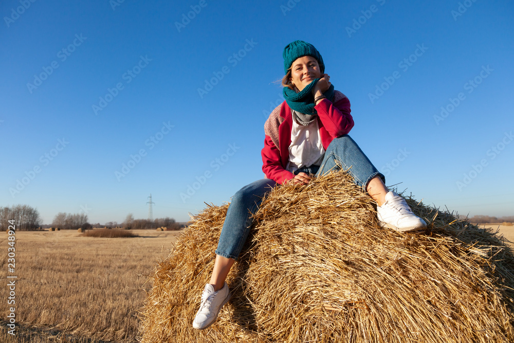 A young woman in a blue knitted hat, pink coat and jeans enjoys nature, sitting on a haystack around blue sky. The concept of livestyle and outdoor recreation in autumn A young woman in a blue knitted hat, pink coat and jeans enjoys nature, sitting on a haystack around blue sky. The concept of livestyle and outdoor recreation in autumn