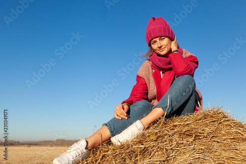 Fashion lifestyle portrait of young trendy woman dressed in pink coat, pink knitted hat  posing, laughing and smiling on a haystack around blue sky.  portrait of joyful woman © Виталий Сова