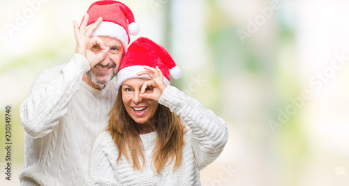 Middle age hispanic couple wearing christmas hat over isolated background doing ok gesture with hand smiling, eye looking through fingers with happy face.