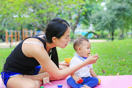 Asian mother feeding food for infant baby boy sitting on pink mattress mat in the garden.