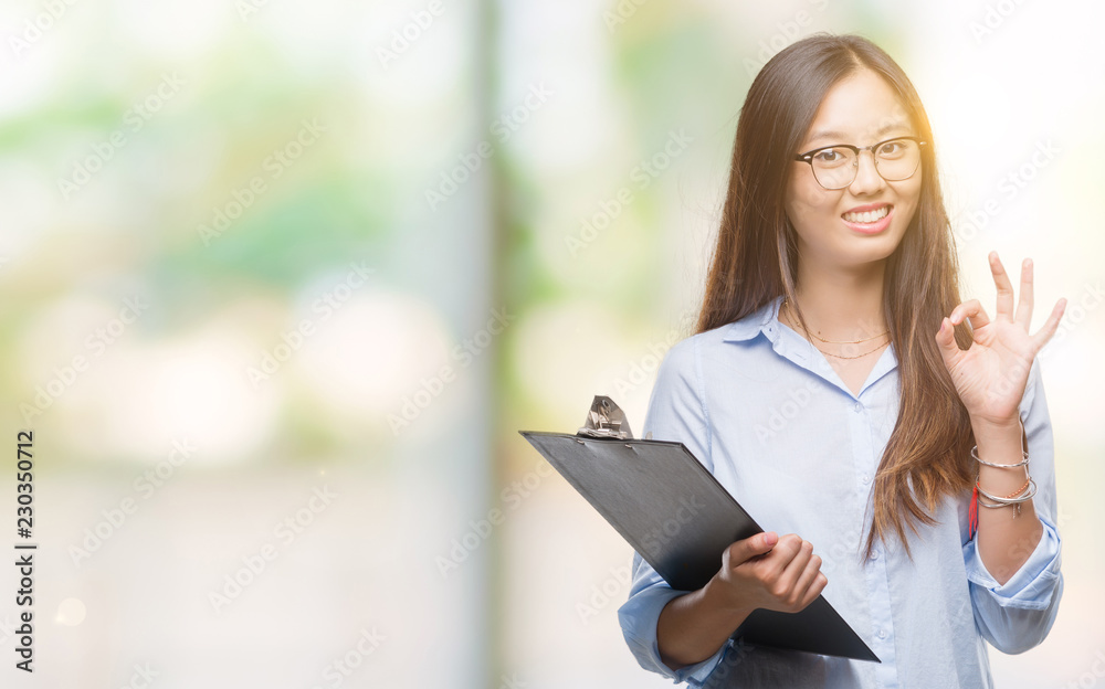 Young asian business woman holding clipboard over isolated background doing ok sign with fingers, excellent symbol Young asian business woman holding clipboard over isolated background doing ok sign with fingers, excellent symbol