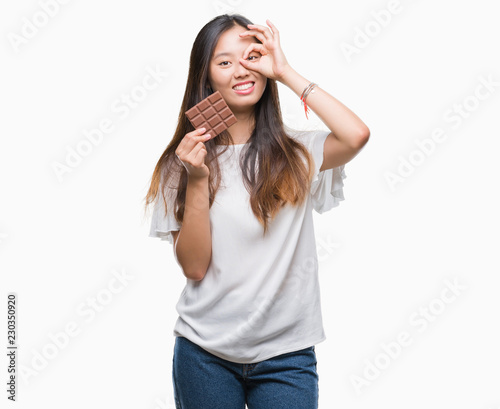Young asian woman eating chocolate bar over isolated background with happy face smiling doing ok sign with hand on eye looking through fingers