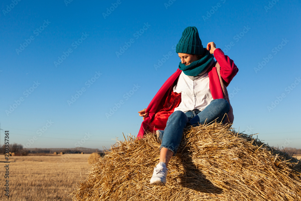 Portrait of a beautiful young model in blue knitted hat and warm clothes have fun, sitting on haystacks in sunny autumn day . Autumn warm photo. Woman smiling and look away, joyful cheerful mood. Portrait of a beautiful young model in blue knitted hat and warm clothes have fun, sitting on haystacks in sunny autumn day . Autumn warm photo. Woman smiling and look away, joyful cheerful mood.
