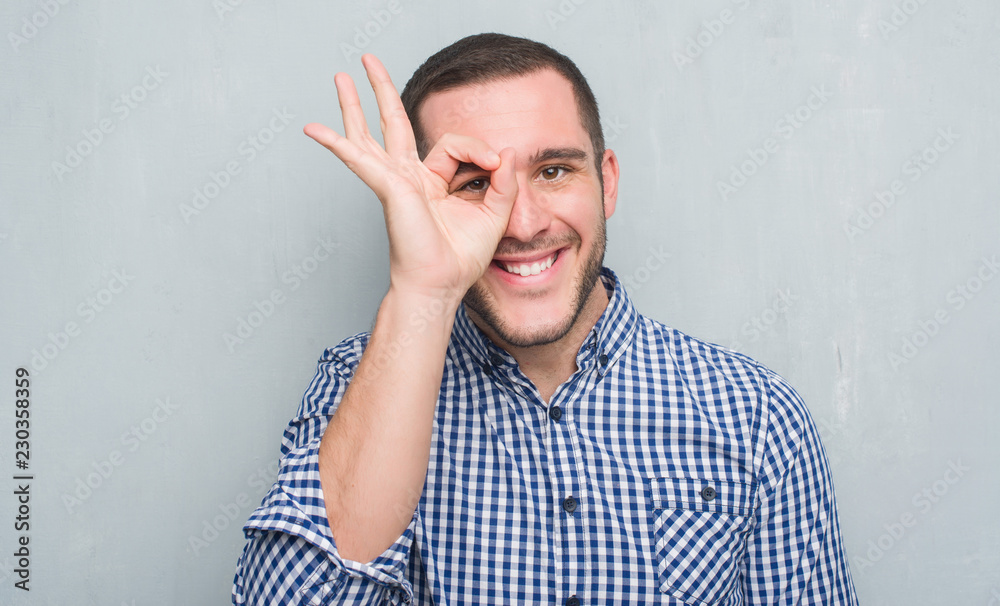 Young caucasian man over grey grunge wall with happy face smiling doing ok sign with hand on eye looking through fingers Young caucasian man over grey grunge wall with happy face smiling doing ok sign with hand on eye looking through fingers
