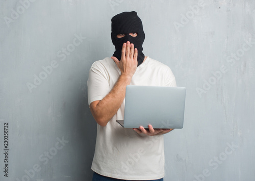 Young caucasian hacker man over grey grunge wall doing cyber attack using laptop cover mouth with hand shocked with shame for mistake, expression of fear, scared in silence, secret concept