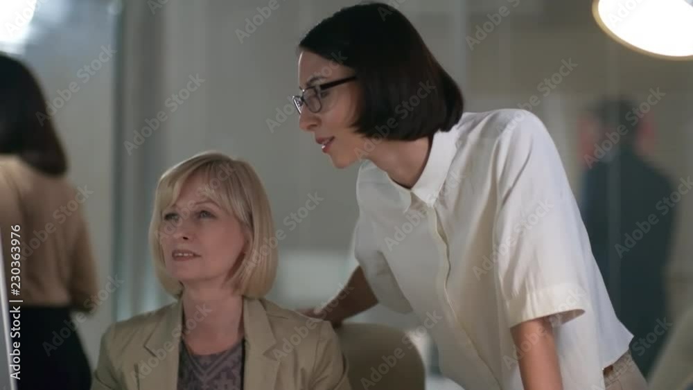 Professional middle aged businesswoman sitting at desk in the office and discussing something on computer screen with young female colleague while working late in the evening Professional middle aged businesswoman sitting at desk in the office and discussing something on computer screen with young female colleague while working late in the evening
