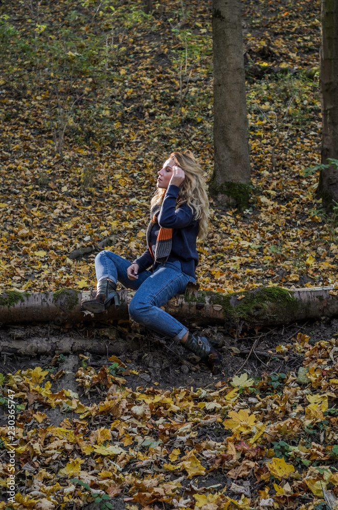 Young beautiful girl with long hair, with a scarf around his neck, sits on a log of fallen tree in the autumn forest among fallen yellow leaves Young beautiful girl with long hair, with a scarf around his neck, sits on a log of fallen tree in the autumn forest among fallen yellow leaves