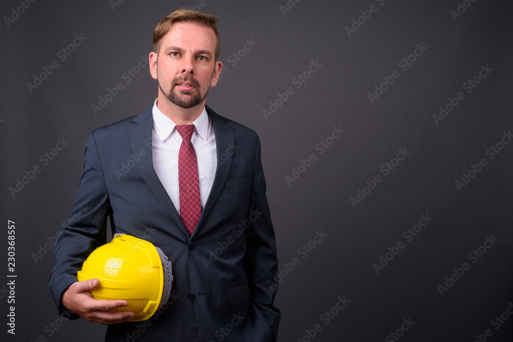 Blond bearded businessman with goatee holding hardhat against gr Blond bearded businessman with goatee holding hardhat against gr