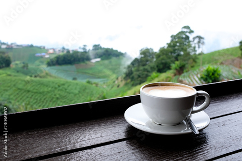A cup of cafe latte coffee in white ceramic cup on wooden table with outdoor green natural mountain view blurred background