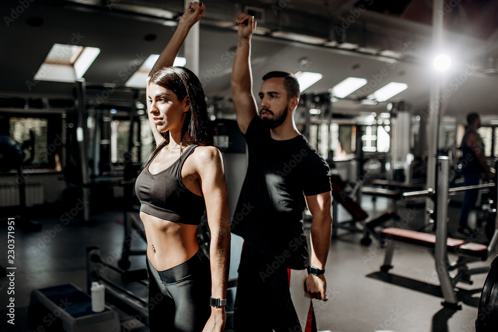 Dark-haired athletic girl dressed in black sport clothes and fitness coach are doing warm up in the gym