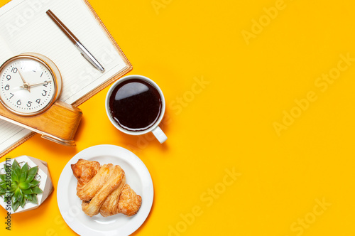 Cup of coffee, open notebook, retro alarm clock, fresh croissant, cactuses succulents on yellow blue minimalistic background, top view Flat Lay with copy space. Empty office desk, Still life, business