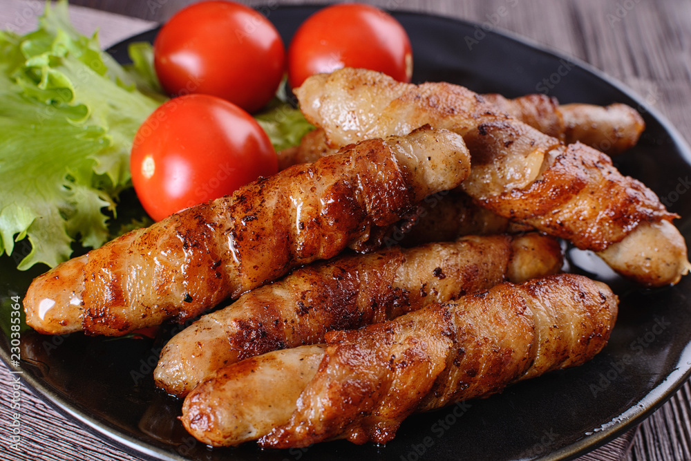 Appetizing and colorful sausages wrapped in bacon and grilled next to tomatoes and lettuce are on a wooden table