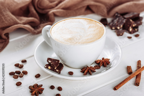 A cup of cappuccino with spices and chocolate pieces on white wooden table. Brown fabric on backdrop