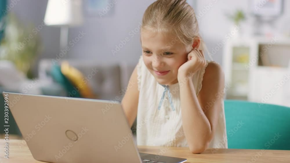 Cute Little Girl Uses Laptop While Sitting at the Desk in the Living Room. Child Has Fun while Browses Through Internet and Watches Cartoons and Funny Videos. Cute Little Girl Uses Laptop While Sitting at the Desk in the Living Room. Child Has Fun while Browses Through Internet and Watches Cartoons and Funny Videos.