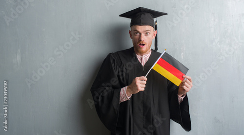 Young redhead man over grey grunge wall wearing graduate uniform holding germany flag scared in shock with a surprise face, afraid and excited with fear expression photo