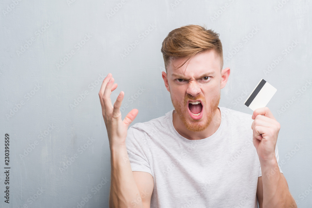 Young redhead man over grey grunge wall holding credit card annoyed and frustrated shouting with anger, crazy and yelling with raised hand, anger concept Young redhead man over grey grunge wall holding credit card annoyed and frustrated shouting with anger, crazy and yelling with raised hand, anger concept