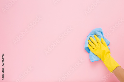 Employee hand in yellow rubber protective glove wiping pastel pink wall from dust with blue dry rag. General or regular cleanup. Commercial cleaning company. Copy space. Empty place for text or logo. photo